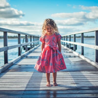 Child Kinjarling Dress cover photos - young girl stood on a jetty. She is wearing a coral pink dress with a bow at the front.